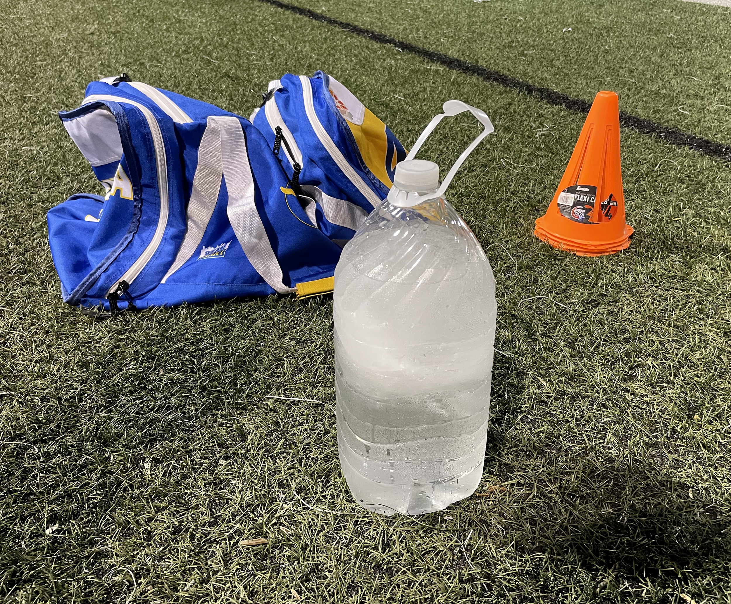 A large water bottle sits on the grass of the football field near a gym bag and orange cones.