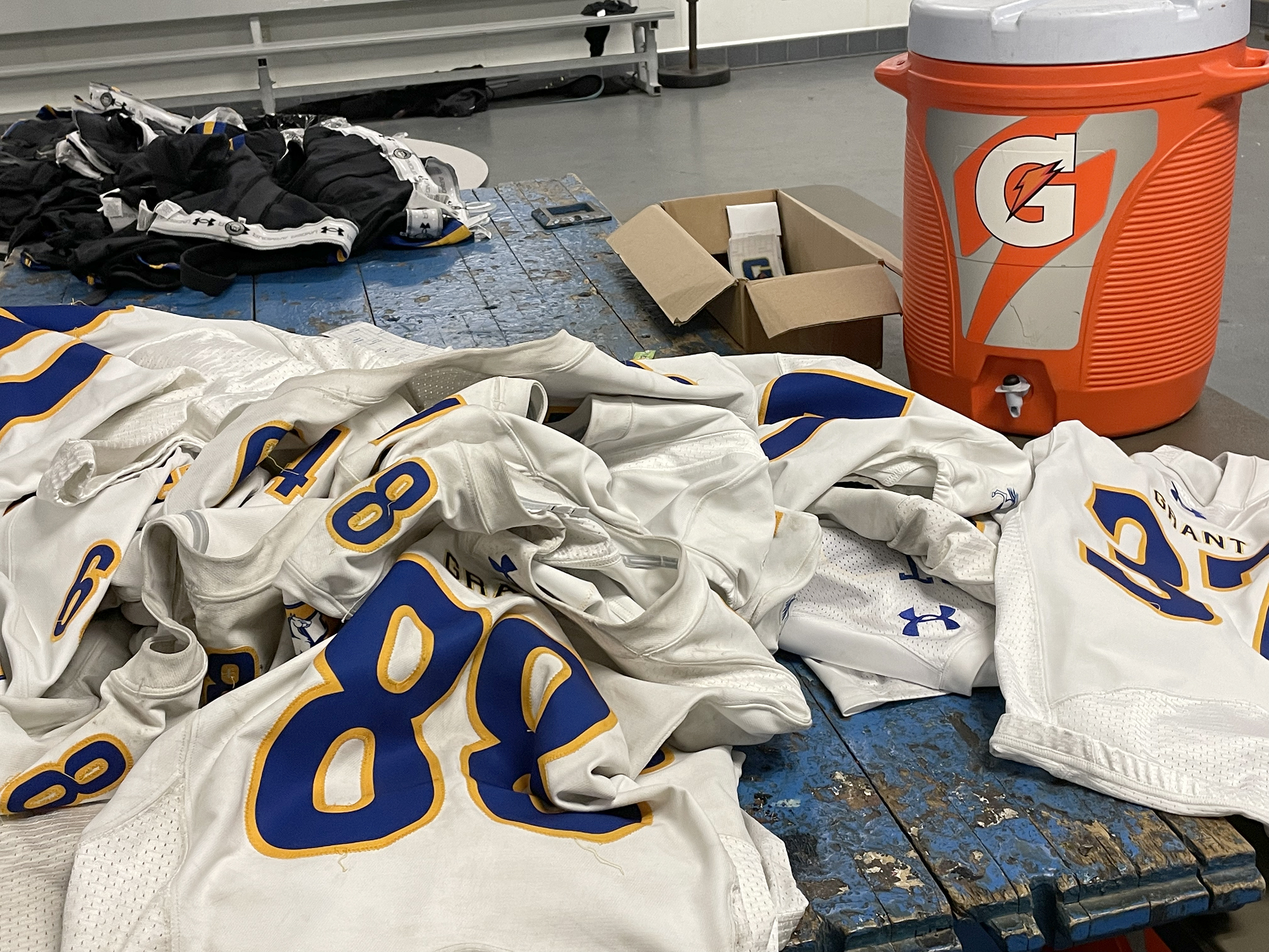 Dirty sport jerseys sit on a blue table in a locker room near a Gatorade cooler.