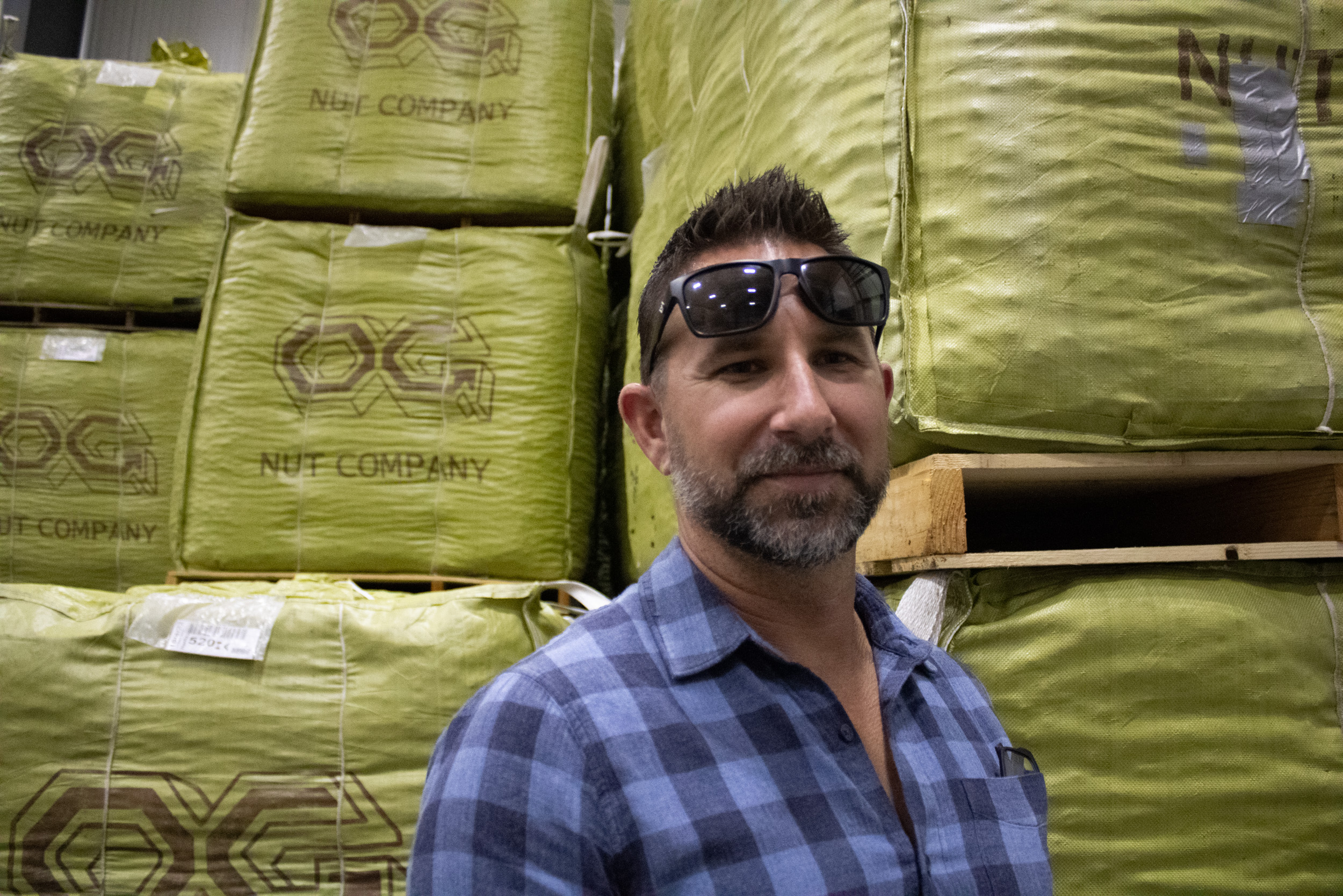 A white man, in a blue flannel shirt, with sunglasses covering his forehead stands in front of three stacks of cubed yellow bags, marked with “OG Nut Company” logos. A single sack  is almost as tall as the man, with three bags are in each stack. 