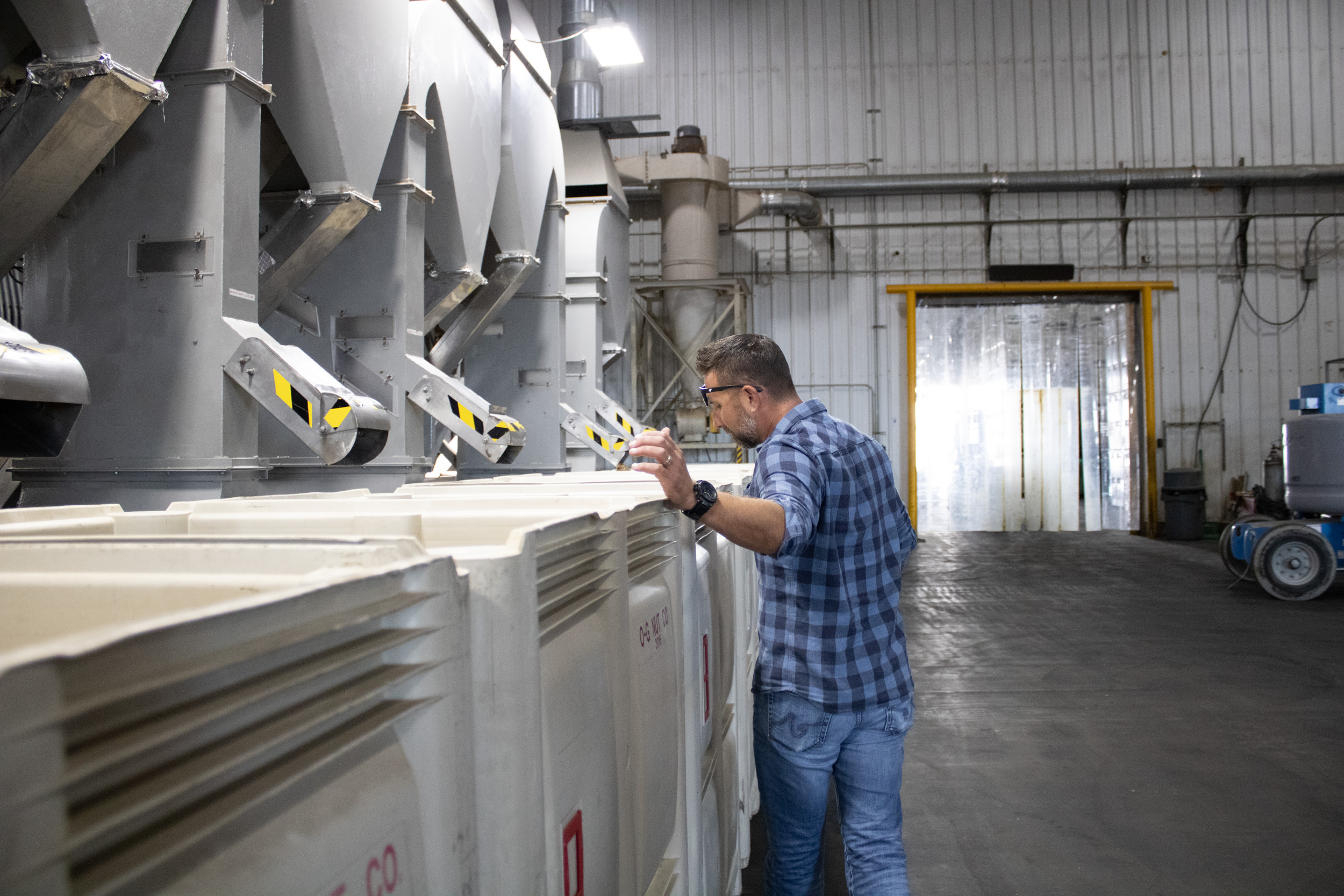 A white man, with short brown hair, wearing a blue shirt and blue jeans, looks into white bins that are almost as tall as he is. A large sorting machine with five sorting shoots sits above the bins, to the left of the man.